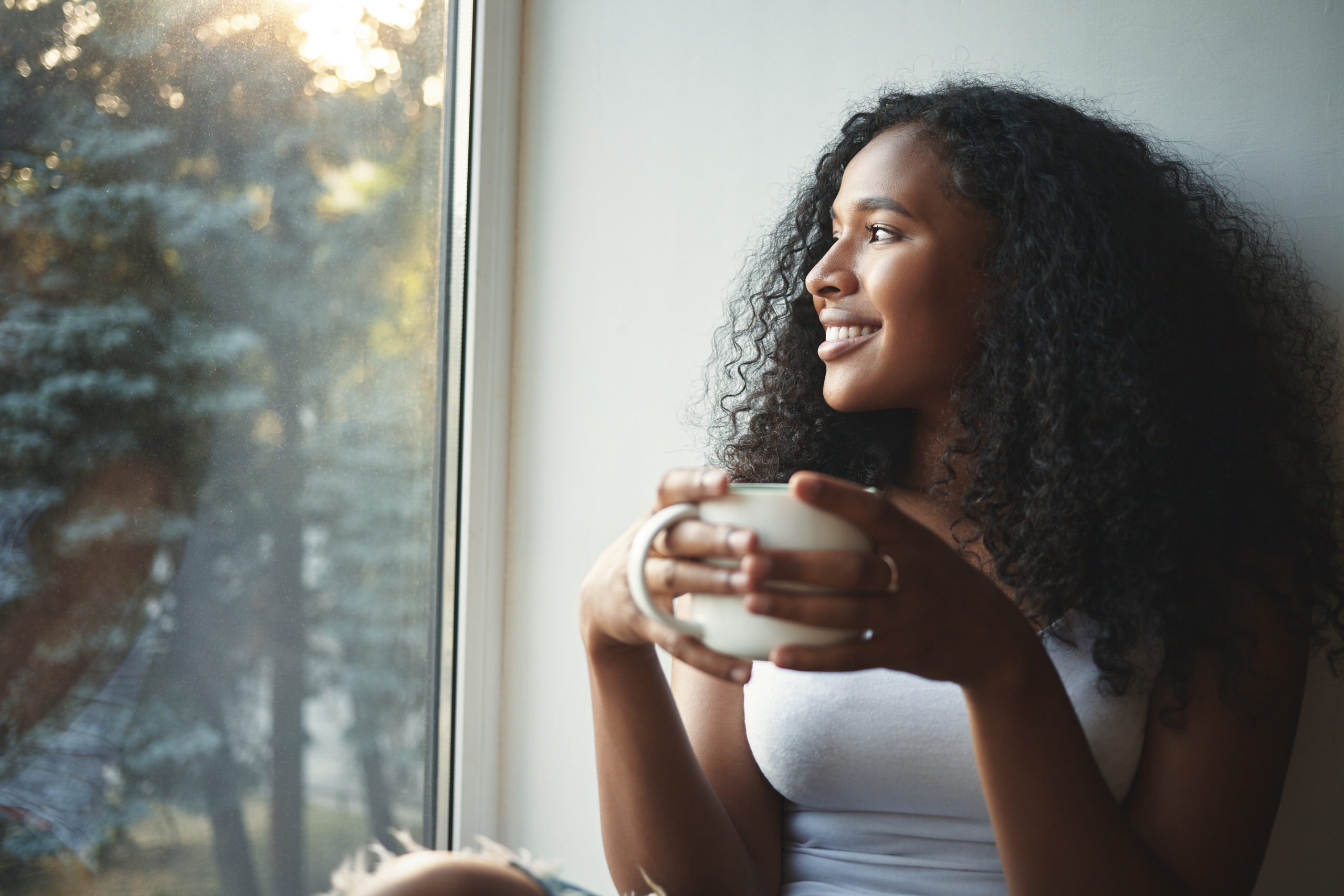 Mulher negra com cabelos escuros segurando uma xícara de café olhando para a janela e refletindo e sorrindo.