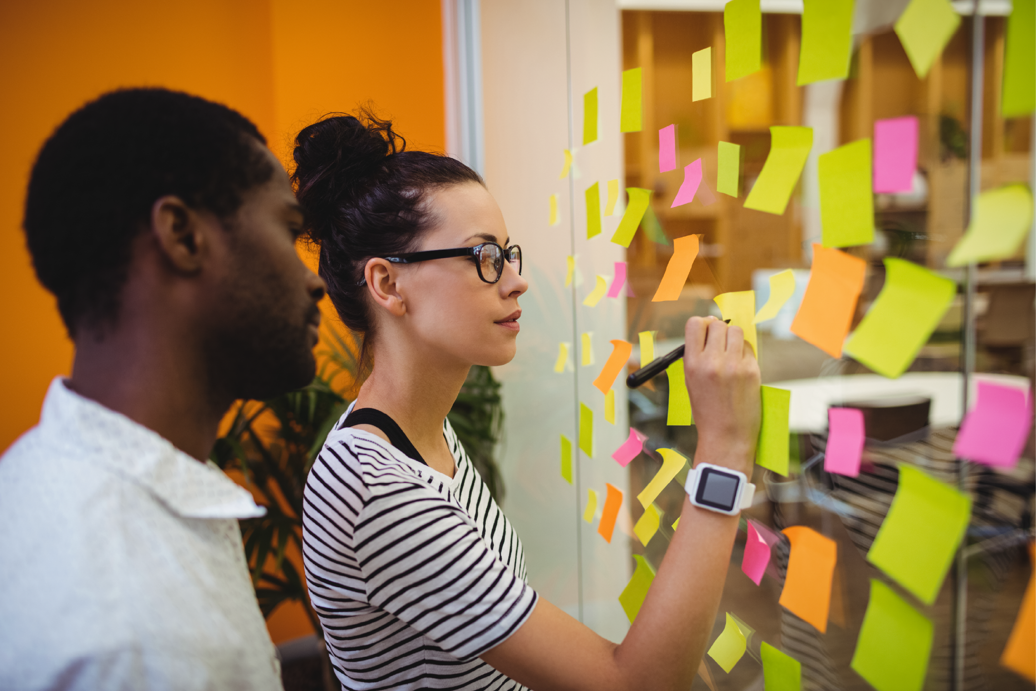 Homem negro e mulher branca escrevendo em post-its numa parede de vidro